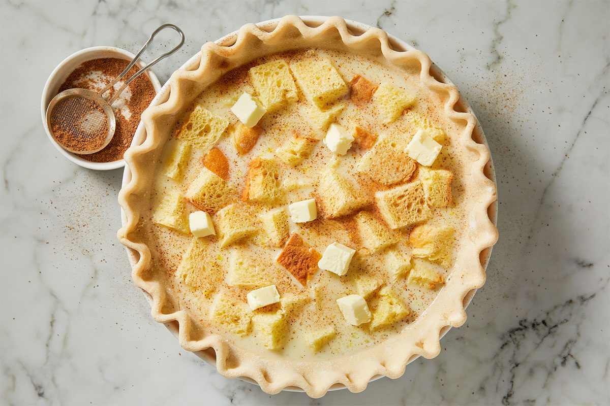 A pie crust filled with a custard mixture, topped with cubed bread and small pieces of butter, is shown on a marble surface next to a bowl of ground spices and a small sifter.
