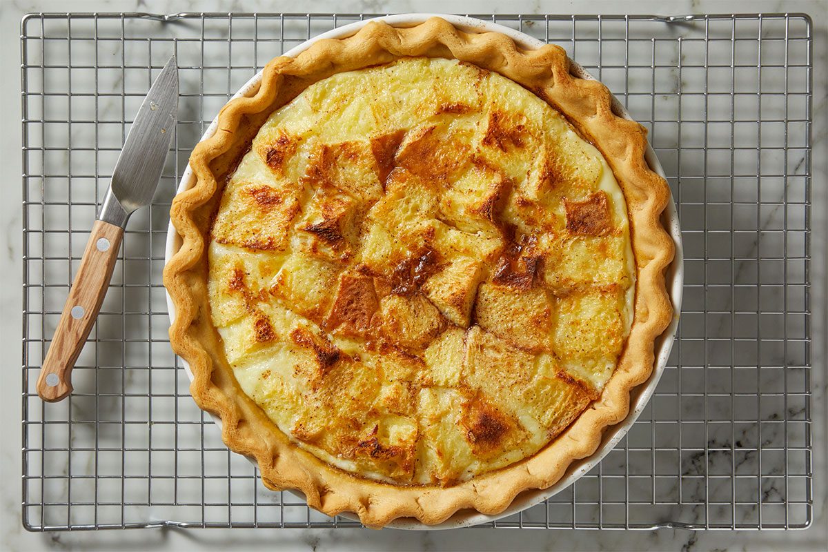 A baked quiche with a golden, flaky crust and browned, cubed bread pieces on top sits on a round white pie dish atop a metal cooling rack, next to a knife with a wooden handle.