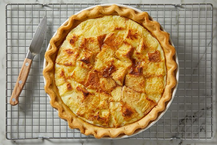 A baked quiche with a golden, flaky crust and browned, cubed bread pieces on top sits on a round white pie dish atop a metal cooling rack, next to a knife with a wooden handle.
