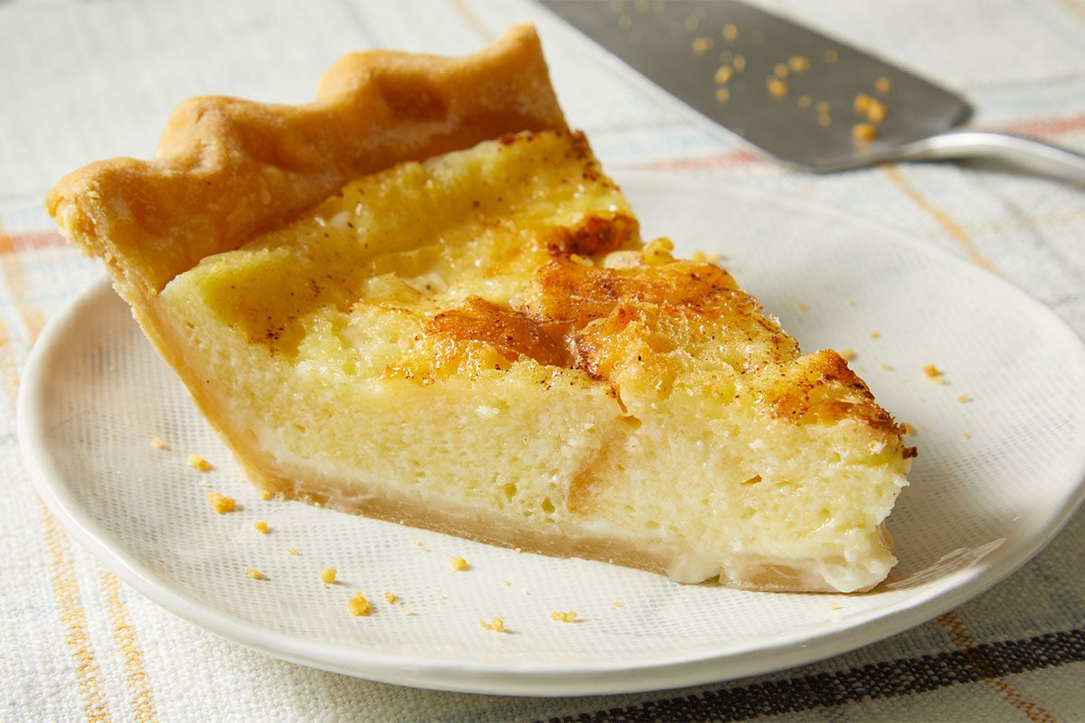 A close-up of a slice of custard pie on a white plate, showing a golden-brown, slightly textured filling and a flaky crust. A pie server is visible in the background on a white tablecloth.