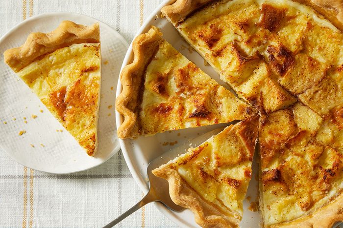 A sliced custard pie with a golden, flaky crust is shown on a plate and a serving dish. One slice is placed separately on a small white plate beside the main pie.