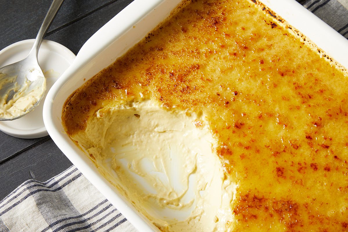 A baking dish filled with creamy crème brûlée, with a portion scooped out, revealing its smooth texture. A spoon with some of the dessert rests on a small plate beside the dish, atop a striped cloth.