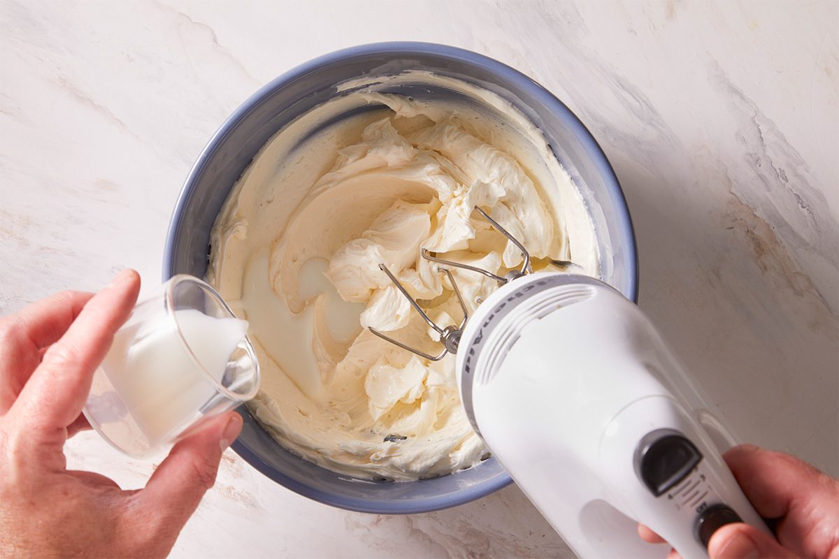 Overhead shot of butter mixture being blended with a hand mixer in a blue mixing bowl as milk is poured in.
