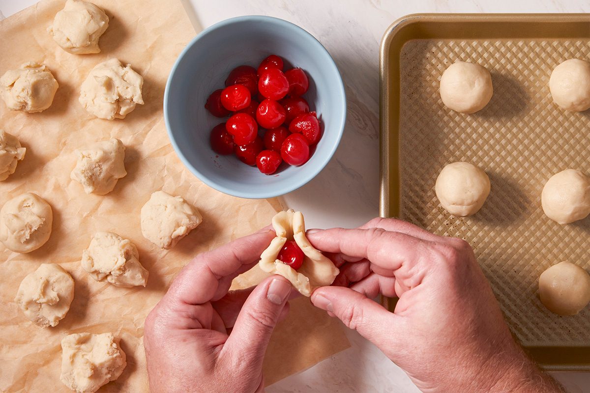 Overhead shot of hands wrapping cookie dough around a red maraschino cherry with dough balls arranged on a baking sheet.