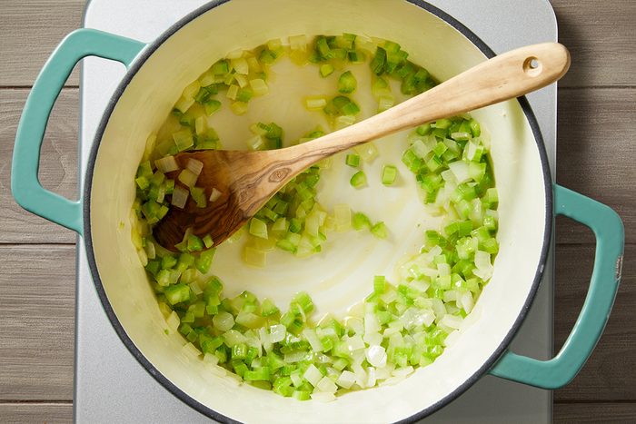 Overhead shot of chopped onions and celery being sautéed in a large light-colored pot with a wooden spoon, as the pot sits on a stovetop
