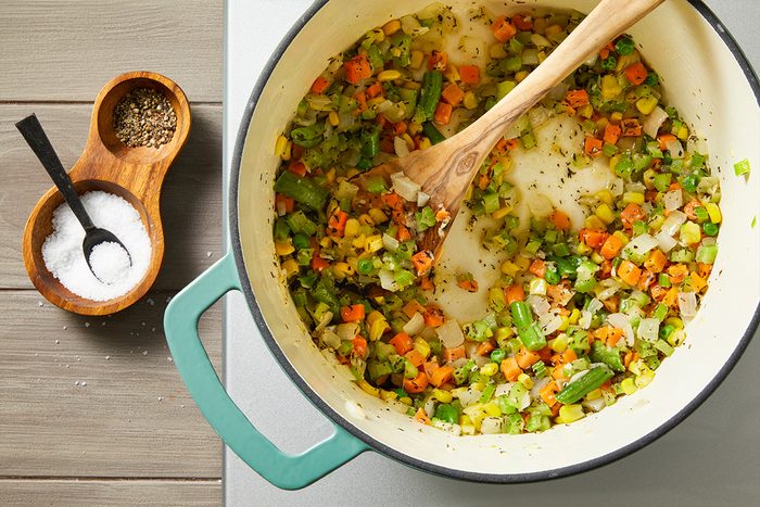 Overhead shot of a pot filled with chopped vegetables carrots, green beans, celery, and onions being stirred with a wooden spoon on a stove, with a small wooden bowl of salt and pepper beside it