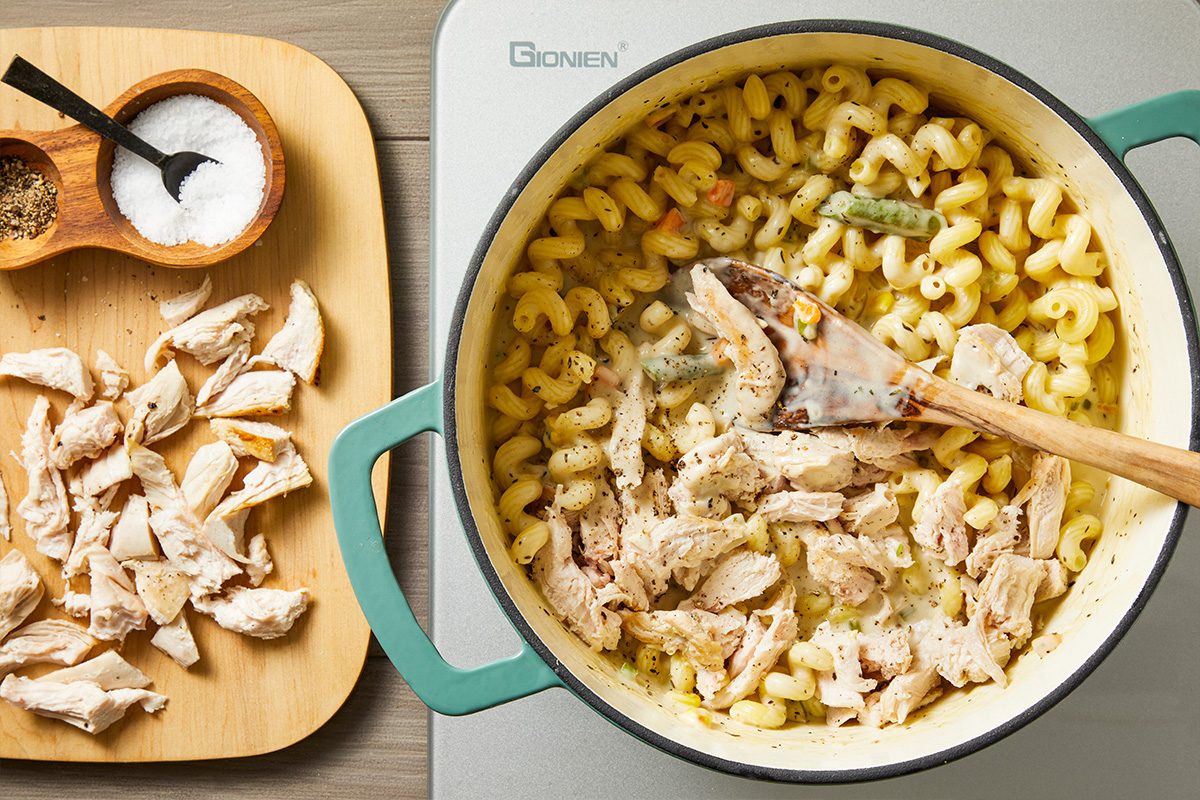 Overhead shot of a pot of creamy pasta with shredded chicken being stirred by a wooden spoon, with a cutting board beside it holding additional shredded chicken and small bowls of salt and pepper