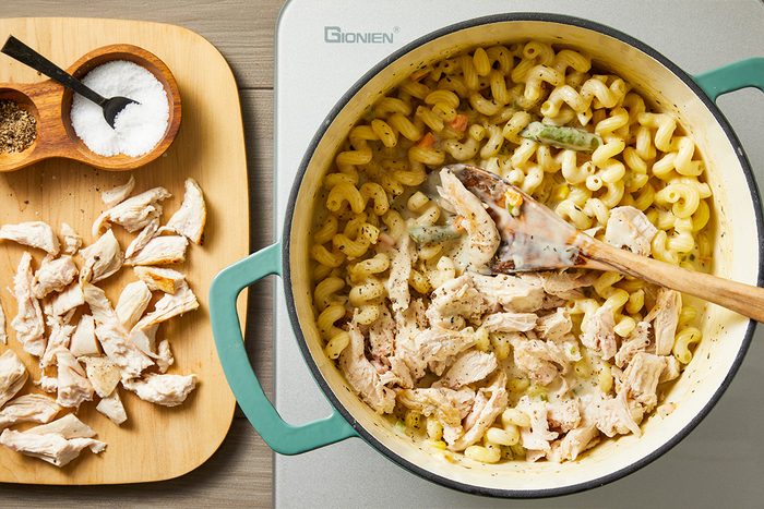 Overhead shot of a pot of creamy pasta with shredded chicken being stirred by a wooden spoon, with a cutting board beside it holding additional shredded chicken and small bowls of salt and pepper