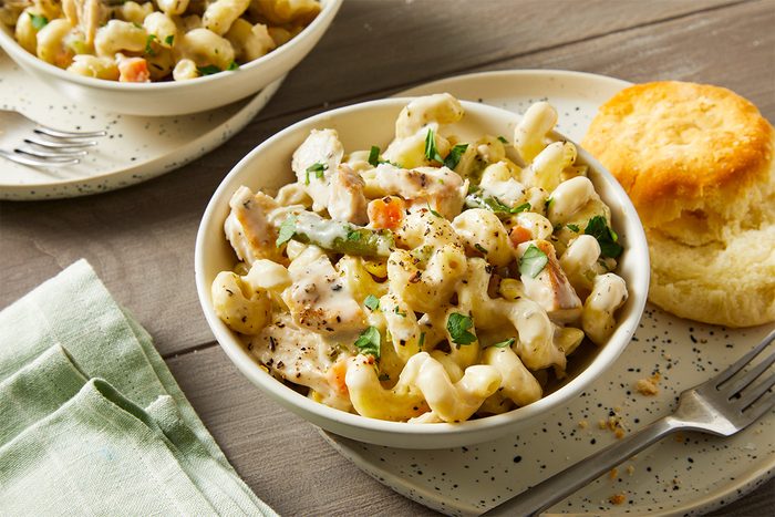 3/4 angle view of a bowl of creamy chicken potpie pasta with curls of pasta, shredded chicken, and mixed vegetables, topped with black pepper and fresh herbs; a flaky biscuit sits on the plate beside the bowl, with a second serving visible in the background