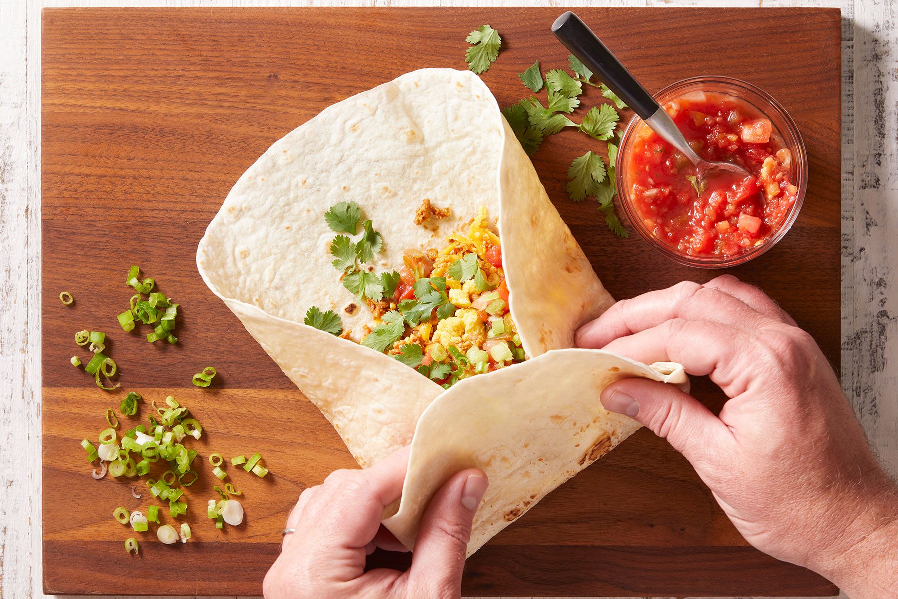 Putting egg and sausage mixture across the center of each tortilla with toppings and folding it into a wrap.