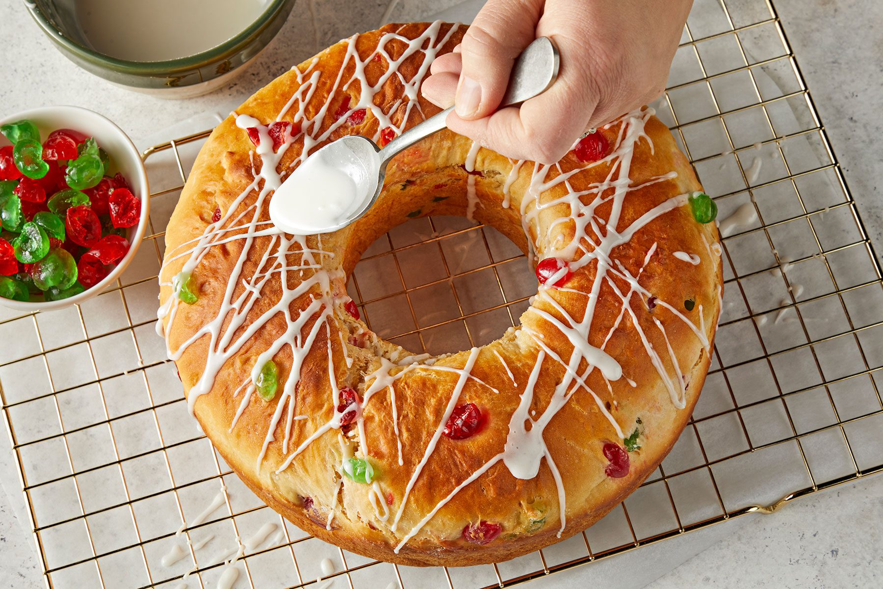 Drizzling glaze over baked bread with some candied fruit placed in a bowl on side.