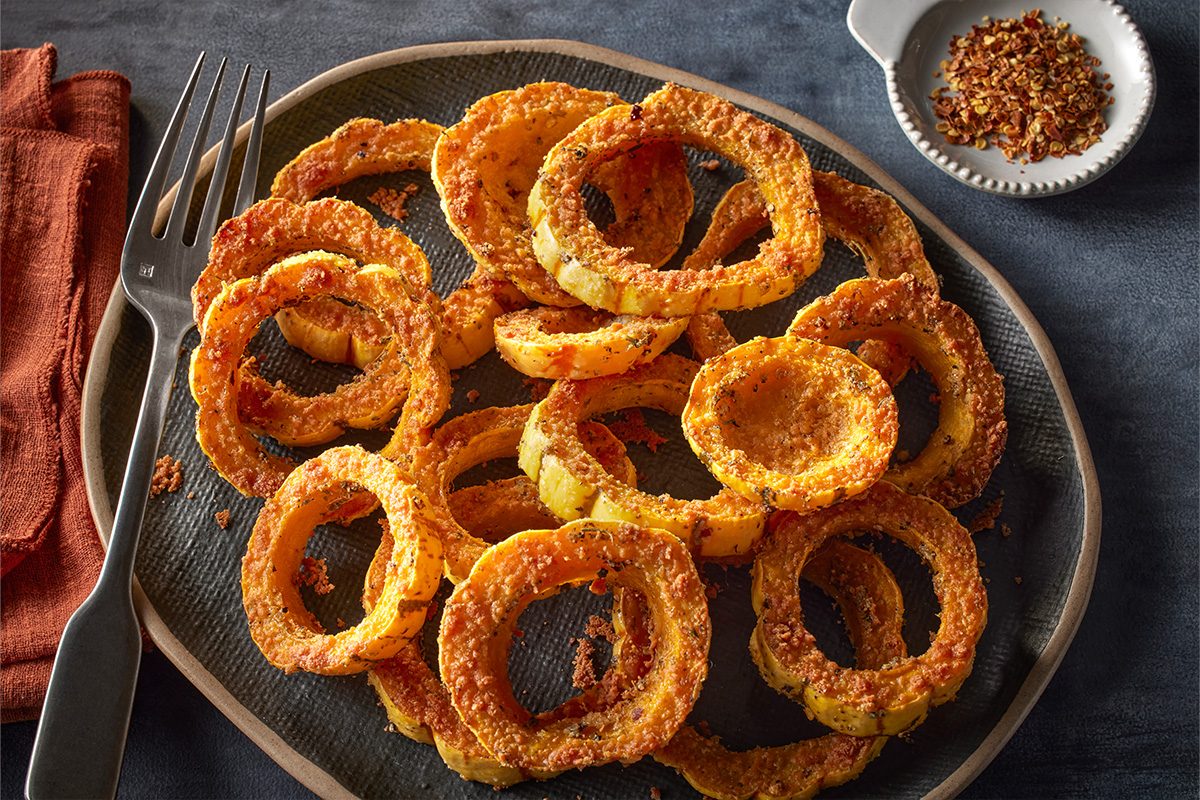 Overhead shot of Crispy Delicata Squash Rings arranged on a dark textured plate, coated with golden seasoning crumbs, with a fork and a small dish of spices on the side;
