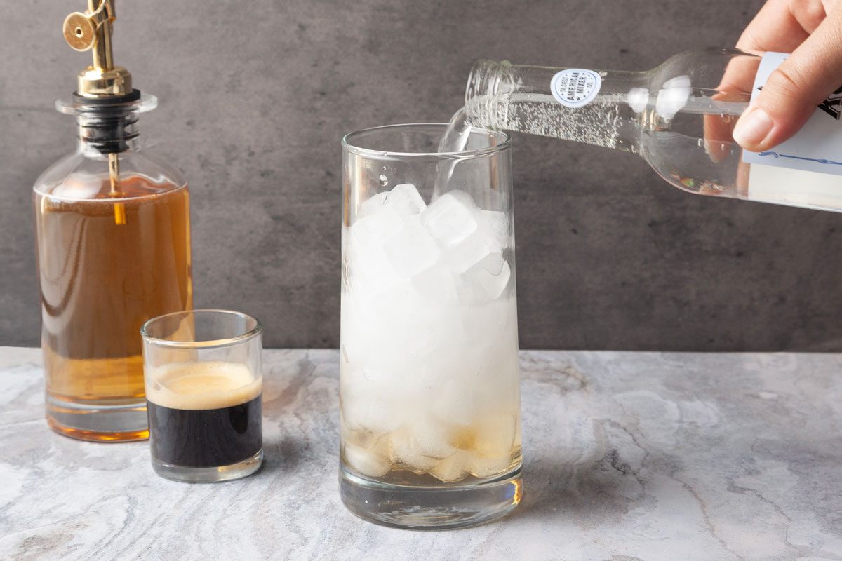 Table view shot of a hand pouring a clear liquid from a bottle into a tall glass filled with ice cubes; Beside it are a short glass of dark liquid and a bottle of amber liquid with a pour spout.