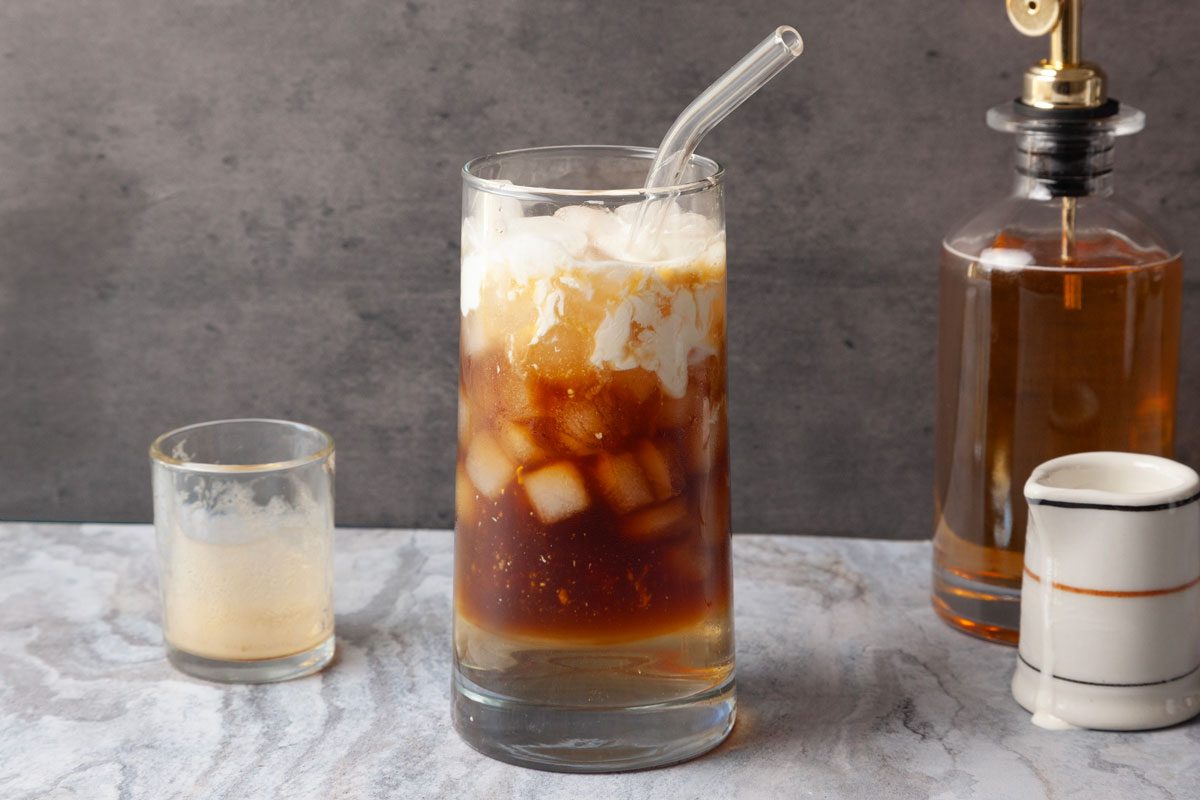 Table view shot of Espresso Cream Soda in a glass with swirling milk and a straw, placed on a marble surface beside a small glass, a pump bottle, and a pitcher.