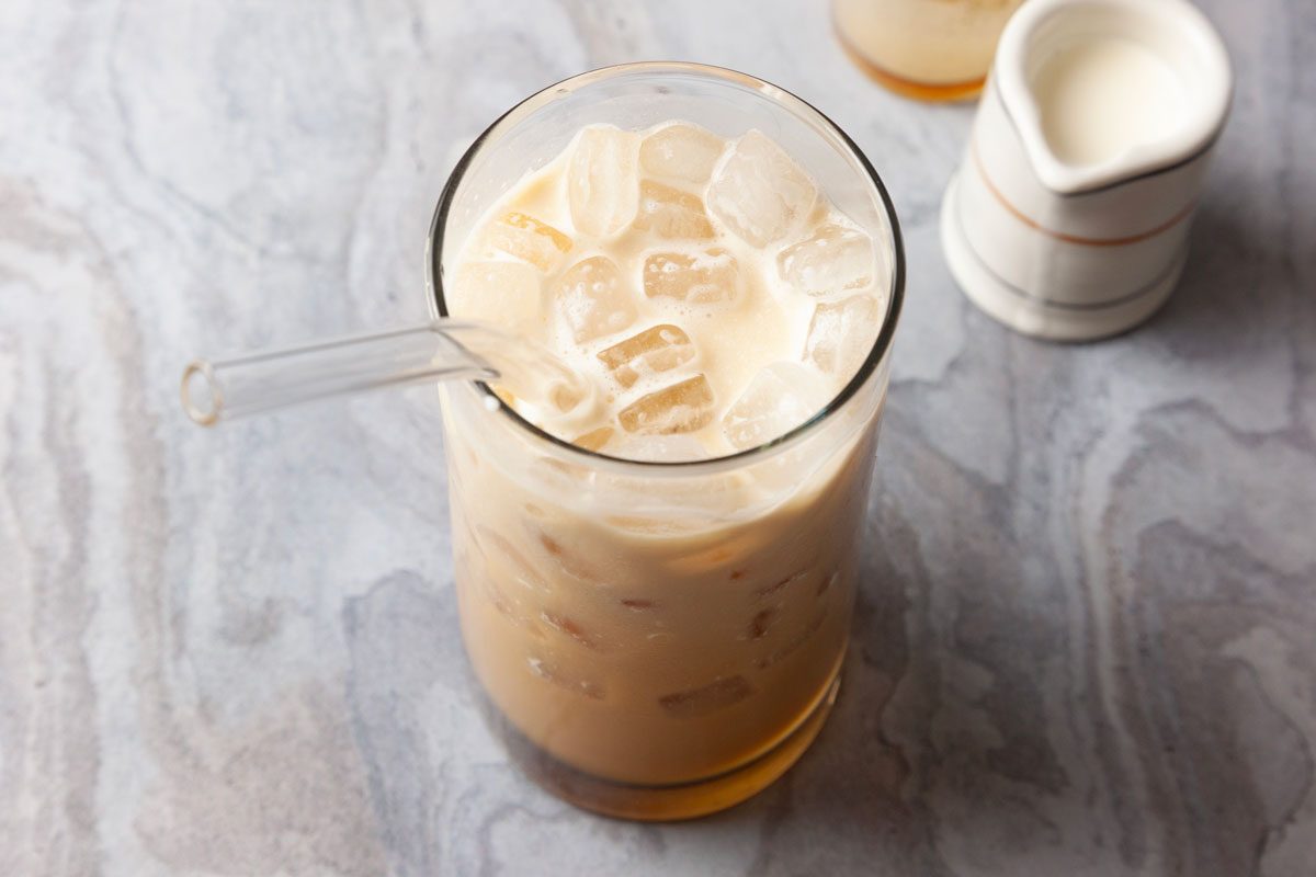 High angle shot of Espresso Cream Soda with milk and ice cubes in a glass with a clear straw, placed on gray marble beside a small white creamer.