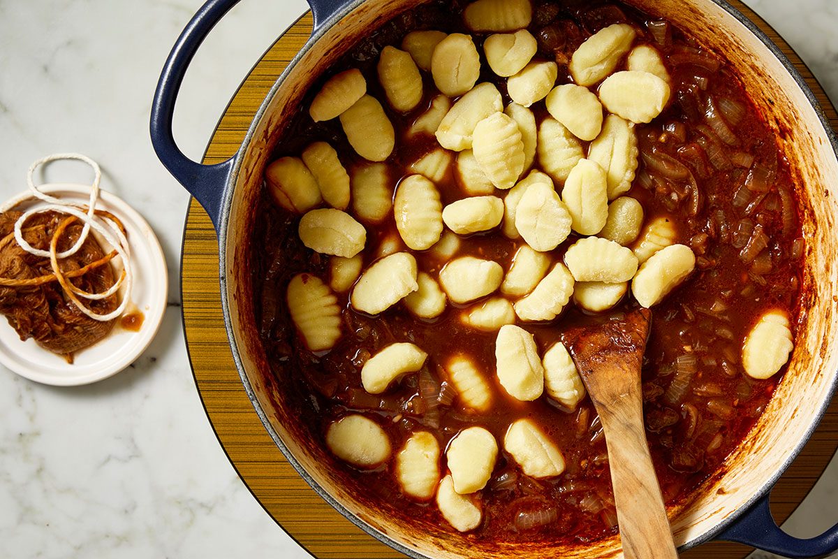 Overhead shot of a pot of tomato-based sauce with gnocchi on top and a wooden spoon inside, placed on a table, with a small dish containing tied herbs or spices nearby.