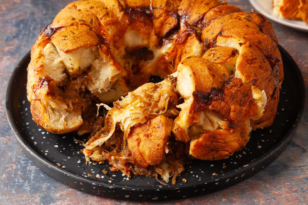 Overhead shot of French Onion Monkey Bread, golden-brown and pull-apart, on a black plate, with servings on two smaller plates revealing its soft, layered texture.