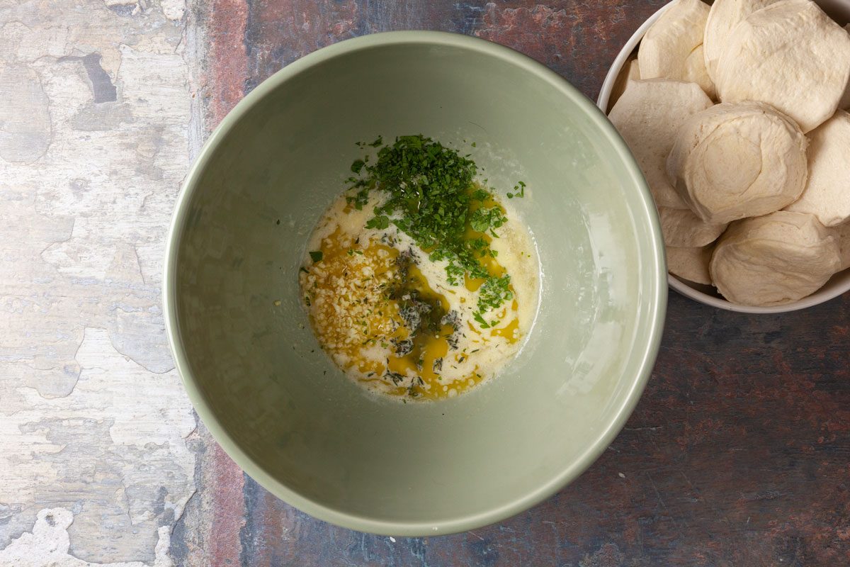 Overhead shot of a green bowl containing melted butter, chopped herbs, and seasoning, placed beside a plate of uncooked biscuit dough rounds on a textured surface.