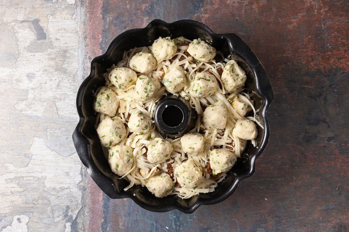 Overhead shot of a bundt pan filled with shredded cheese and small dough balls sprinkled with herbs, ready to be baked, placed on a rustic, textured surface.