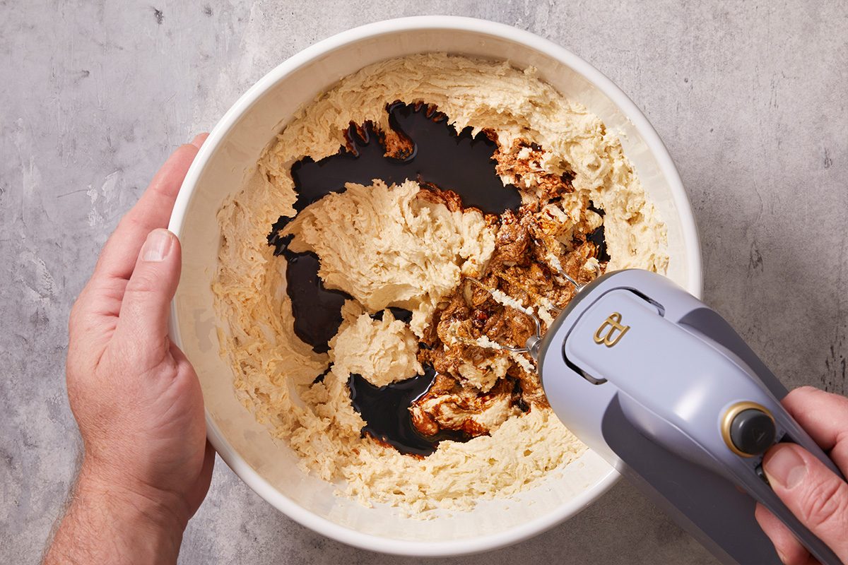 Overhead shot of a hand mixer blending cookie dough with added molasses in a white bowl.