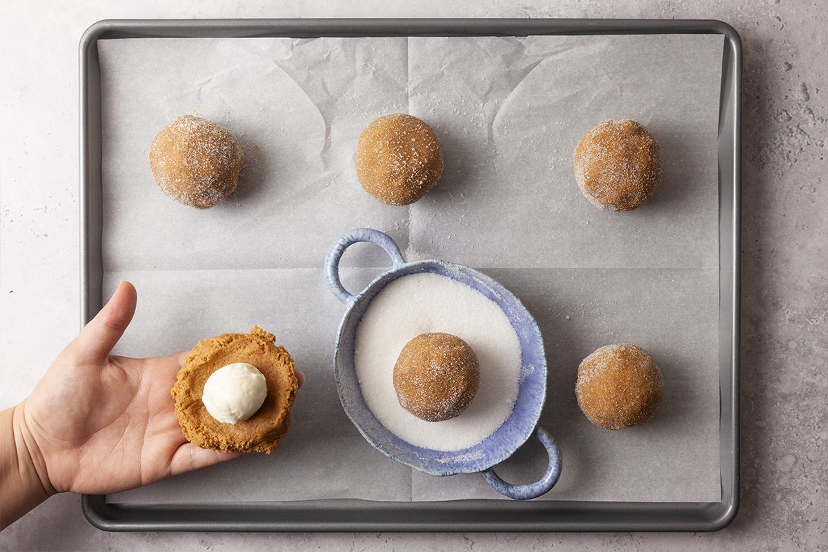 Overhead shot of a hand holding a flattened dough ball filled with cream cheese, positioned above a parchment-lined tray; Five sugar-coated dough balls rest on the tray, and a small bowl of sugar sits in the center;