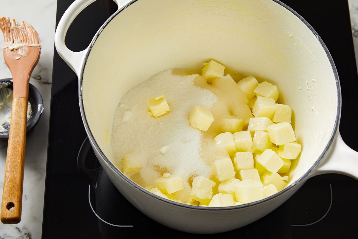 Overhead shot of butter cubes melting with sugar in a white saucepan on the stovetop, wooden spoon nearby.