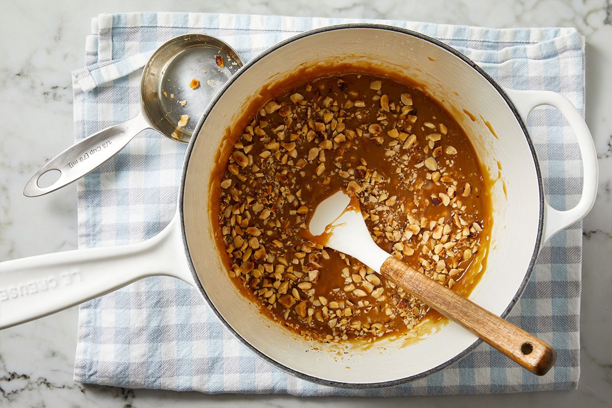 Overhead shot of a saucepan with golden toffee mixture stirred with a wooden spoon, resting on a checkered towel.