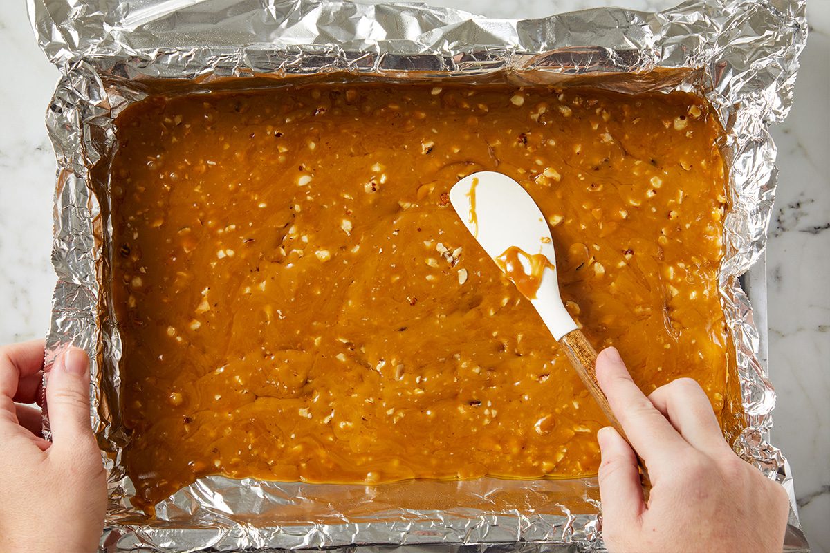 Overhead shot of melted toffee being spread evenly in a foil-lined baking pan using a white spatula.
