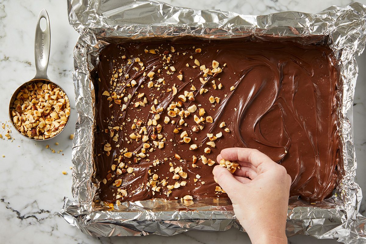 Overhead shot of chopped nuts being sprinkled over melted chocolate spread atop toffee in the pan.