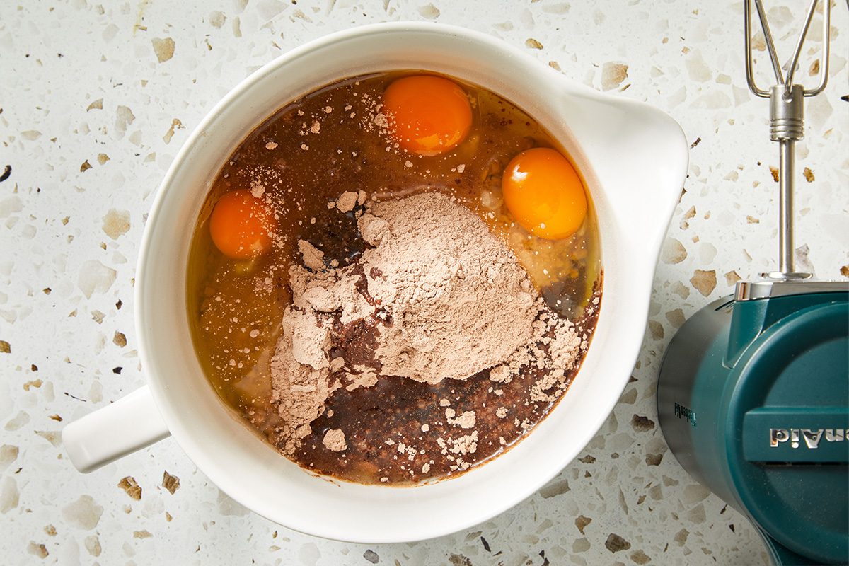 Eggs, cocoa powder, and flour ready to be mixed in a white bowl with a hand mixer nearby.