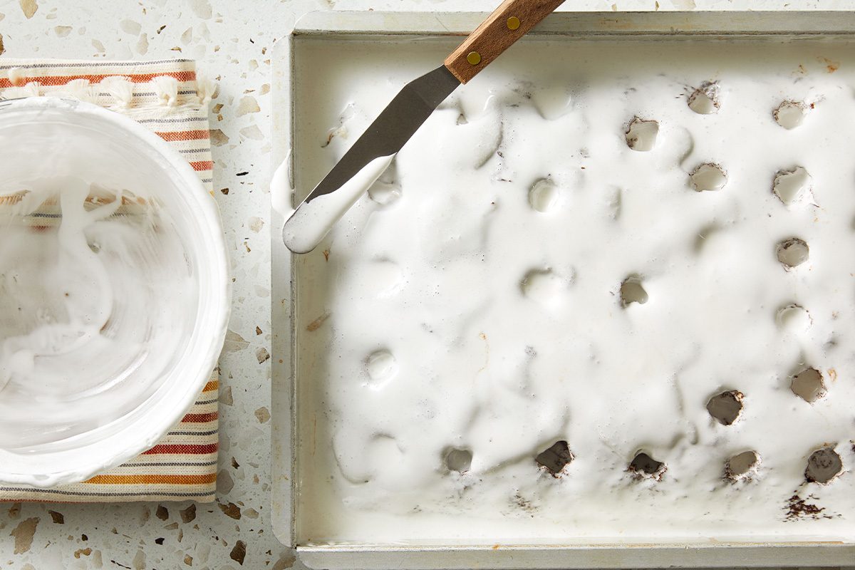 Top-down view of the poke cake covered with a thick layer of whipped cream, with an offset spatula resting nearby.