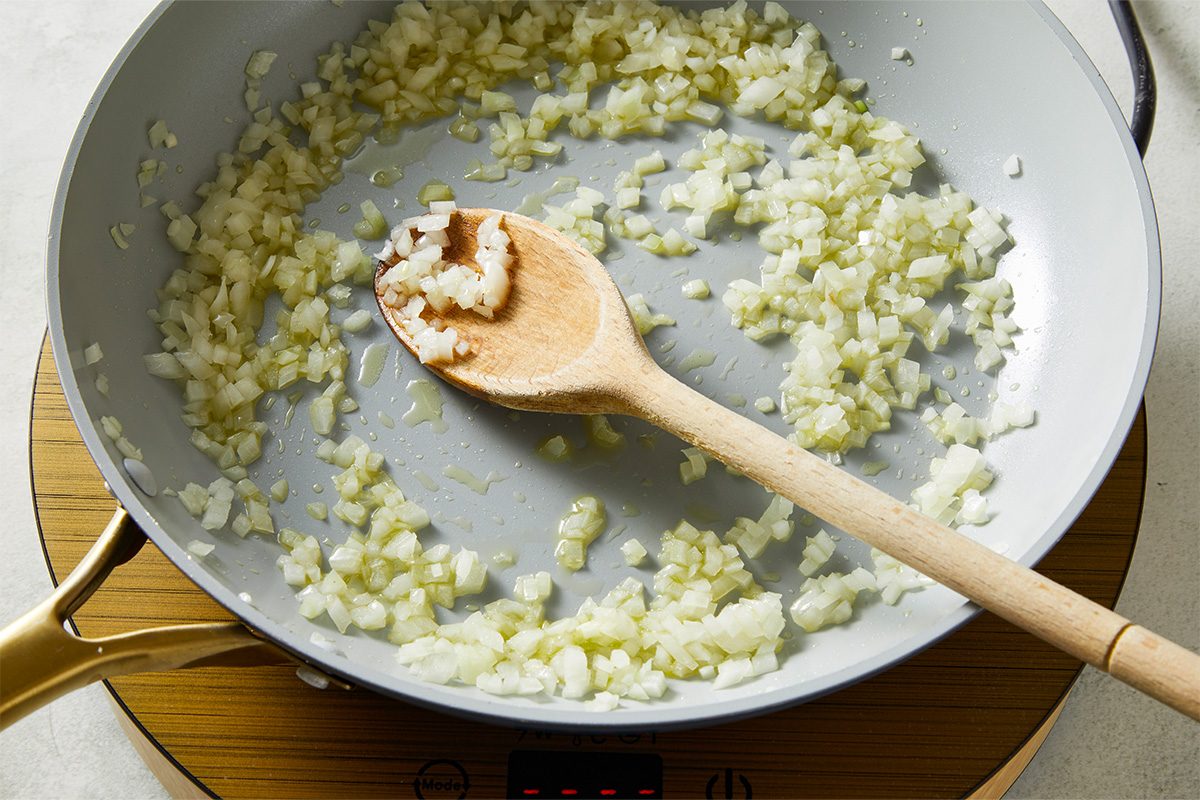 Closeup shot of chopped onions being sautéed in a light-colored frying pan on an electric stove, stirred with a wooden spoon as they begin to turn translucent;