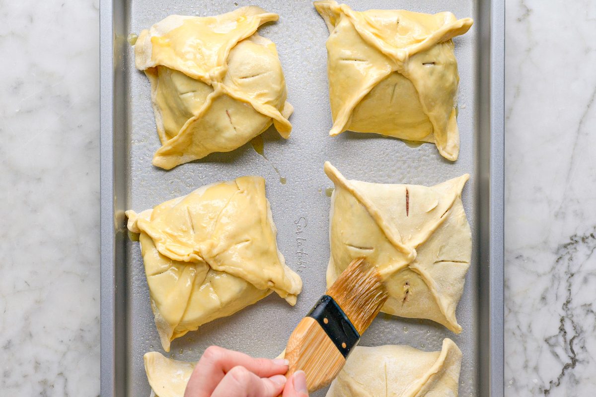 Overhead shot of a hand using a pastry brush to apply egg wash to folded dough parcels on a baking sheet, preparing them for baking on a marble countertop;