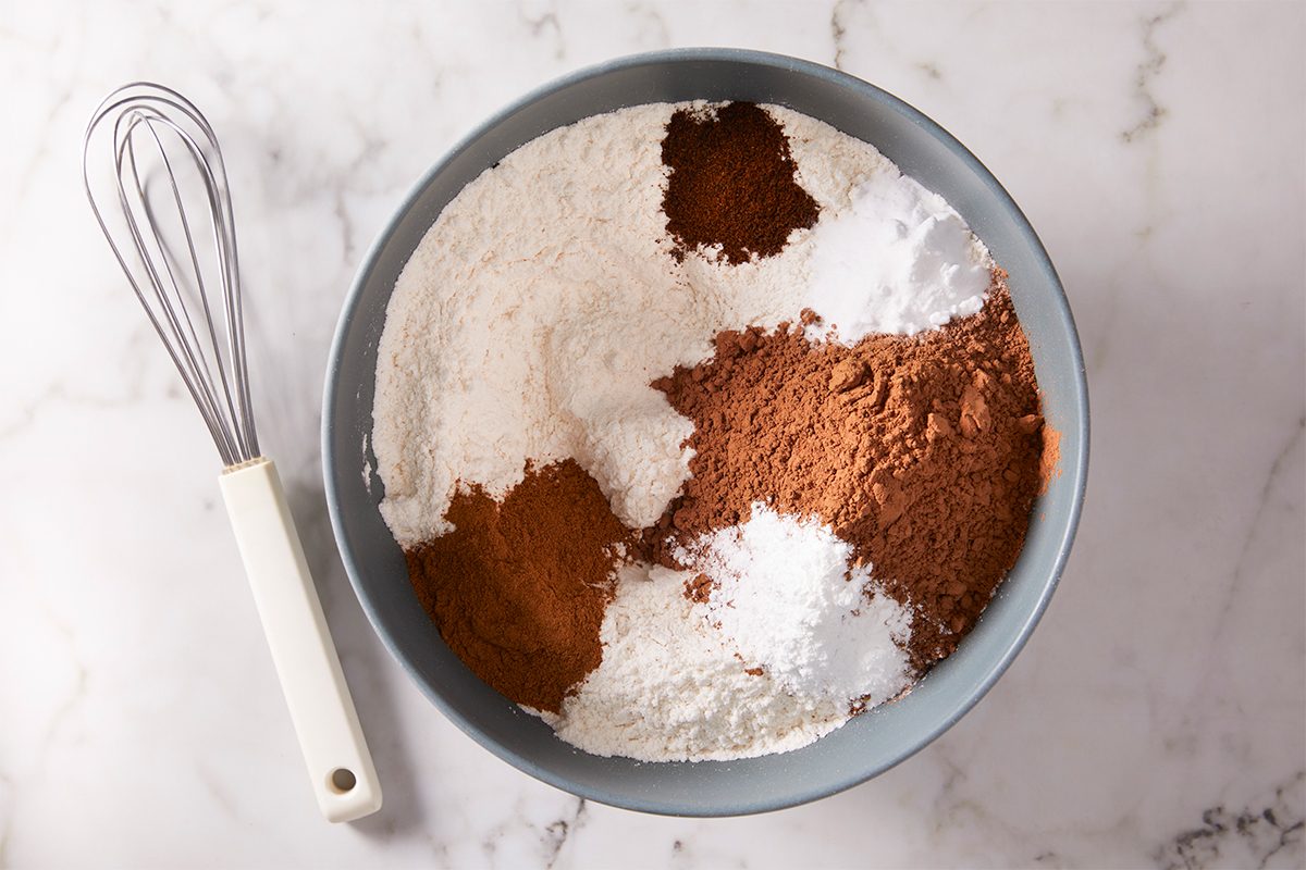 A gray bowl filled with flour, cocoa powder, baking soda, baking powder, and spices sits on a marble surface next to a metal whisk with a white handle.