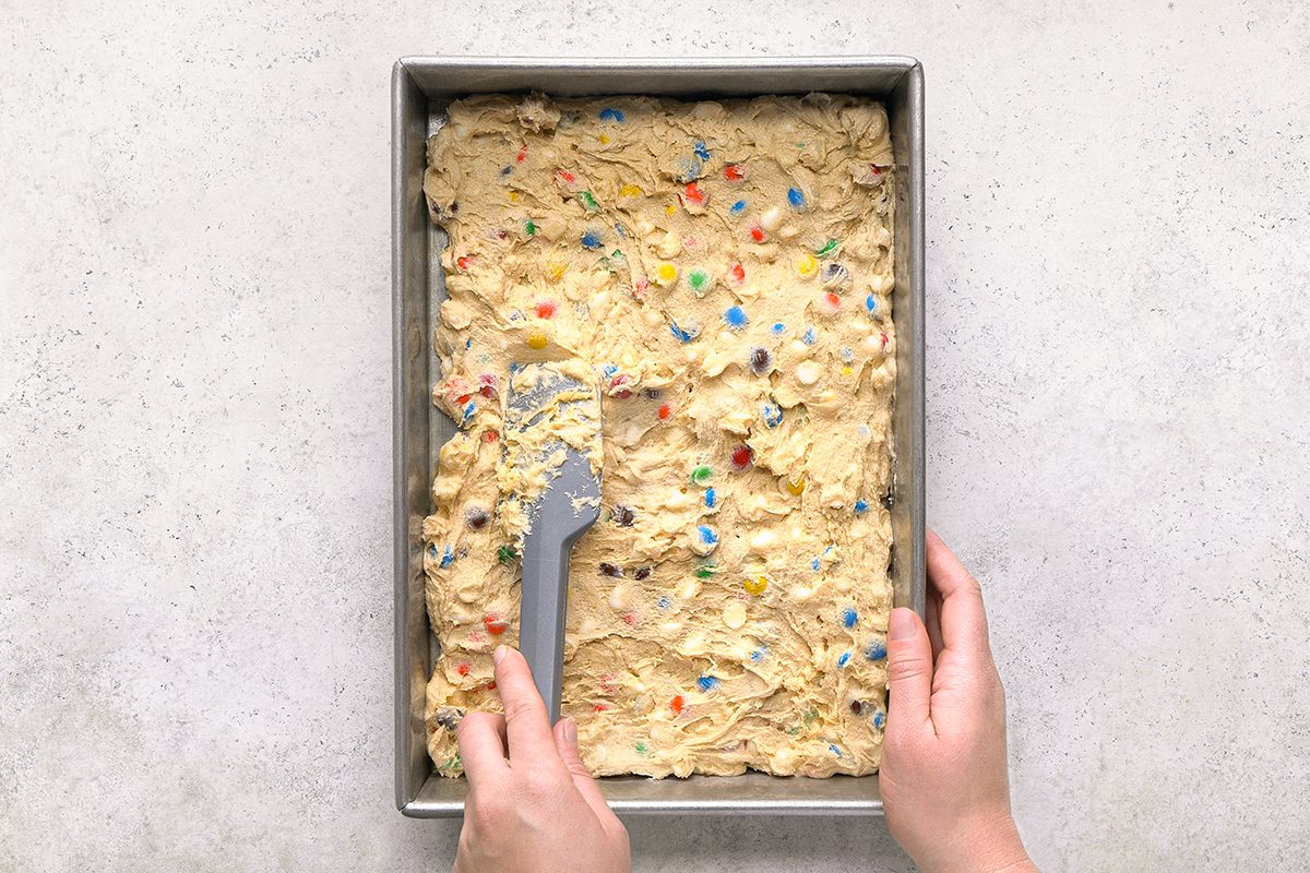 A person spreads colorful candy-studded cookie dough evenly in a rectangular baking pan using a spatula, seen from above on a light gray surface.