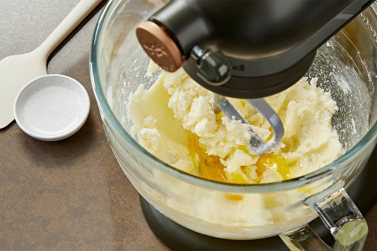 Overhead shot of a stand mixer with a glass bowl creaming butter and sugar, with a cracked egg added, and a white spatula and small empty dish on the brown countertop nearby;