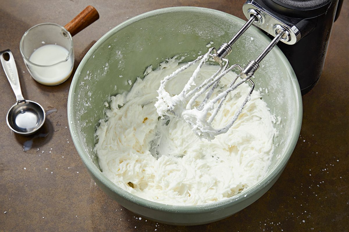Overhead shot of a mixing bowl filled with thick whipped cream on a counter, with an electric hand mixer resting on the bowl. Nearby are a measuring spoon and a small cup containing liquid;