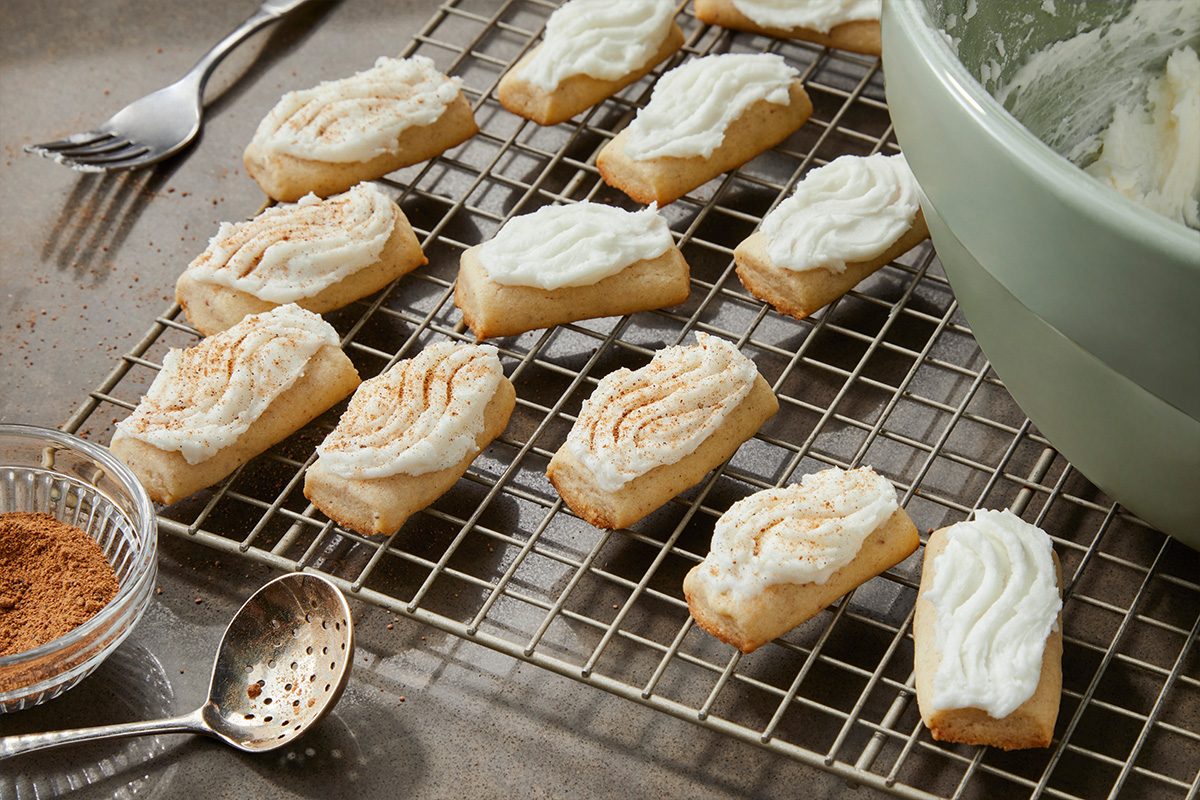3/4 angle view shot of rectangular cookies with white icing on a cooling rack, some sprinkled with cinnamon, with a green bowl of cream, a small dish of cinnamon, and a spoon nearby on a gray surface;
