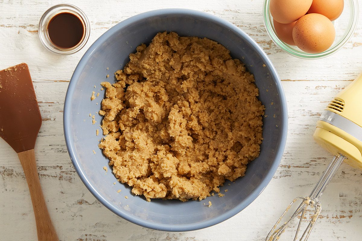 Overhead shot of a mixing bowl with cookie dough in progress, surrounded by eggs and vanilla extract on a marble surface.