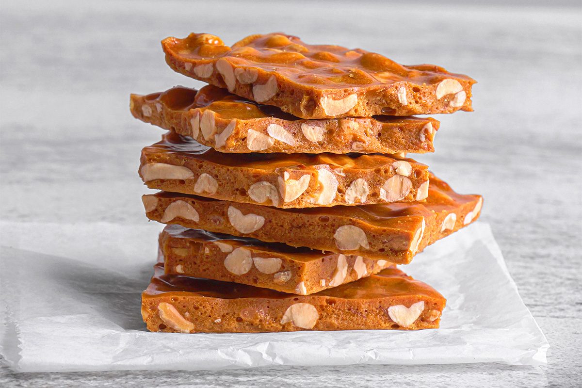 A stack of golden-brown peanut brittle pieces with visible peanuts, resting on white parchment paper against a light gray background.