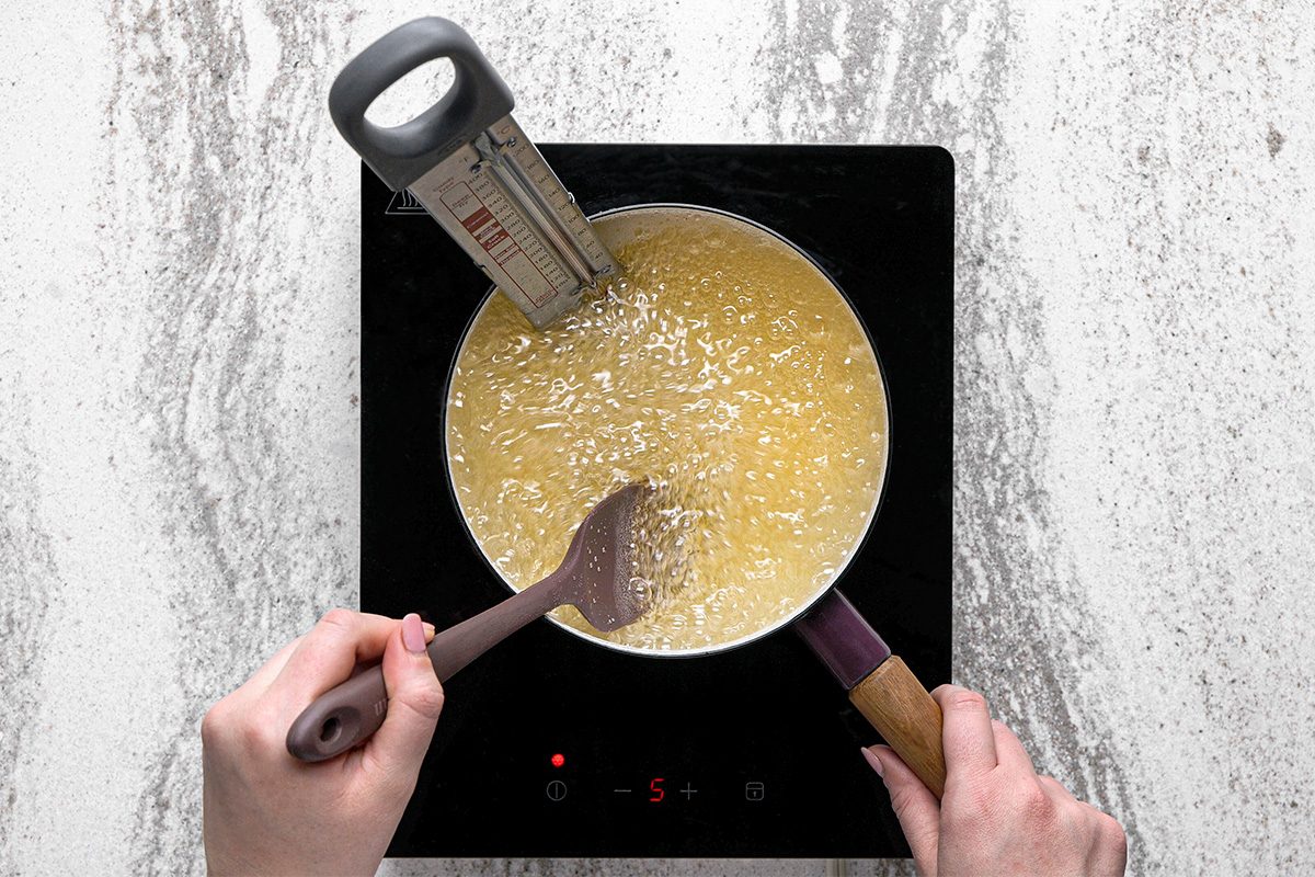 A person stirs oil with a slotted spoon in a pot on an induction cooktop, using a thermometer to measure the temperature for frying.