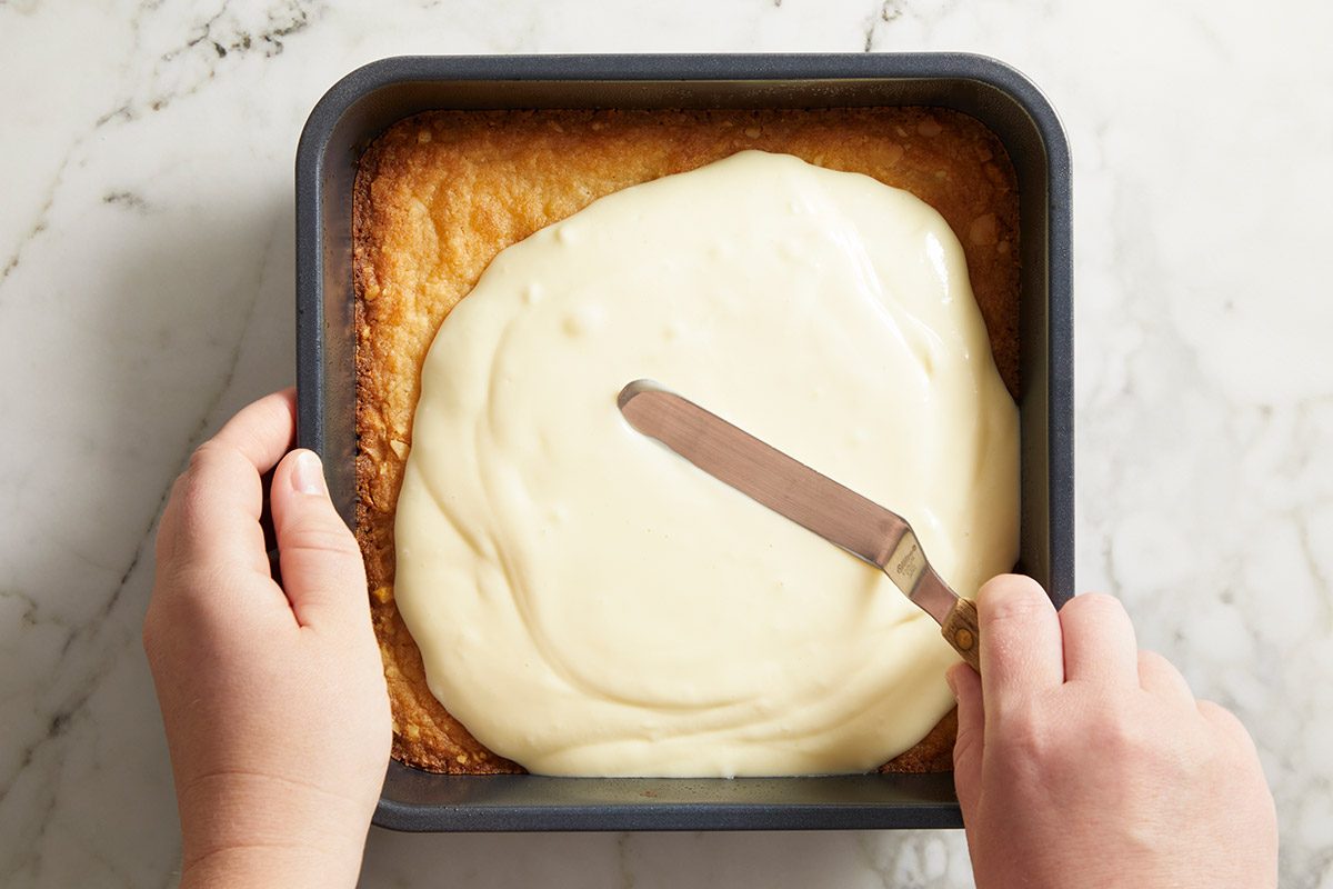 Overhead shot of hands spreading the cream cheese custard layer over the baked crust in a square baking pan.