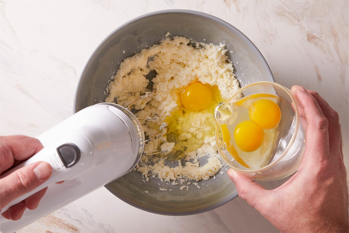 Overhead shot of butter and sugar mixture being blended in a gray bowl while eggs are poured in.
