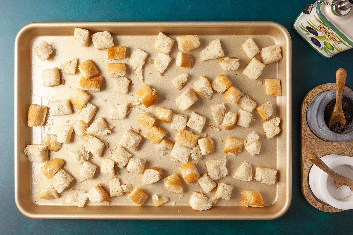 Overhead shot of cubed bread pieces spread out on a baking sheet, ready for baking, with a container of olive oil, a small bowl of pepper, and a small bowl of salt placed on a wooden tray nearby.