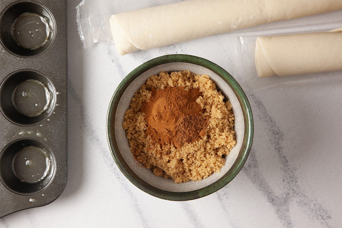 Overhead shot of a bowl of brown sugar mixed with ground cinnamon on a countertop, beside two rolled pieces of dough and a partially visible muffin tin
