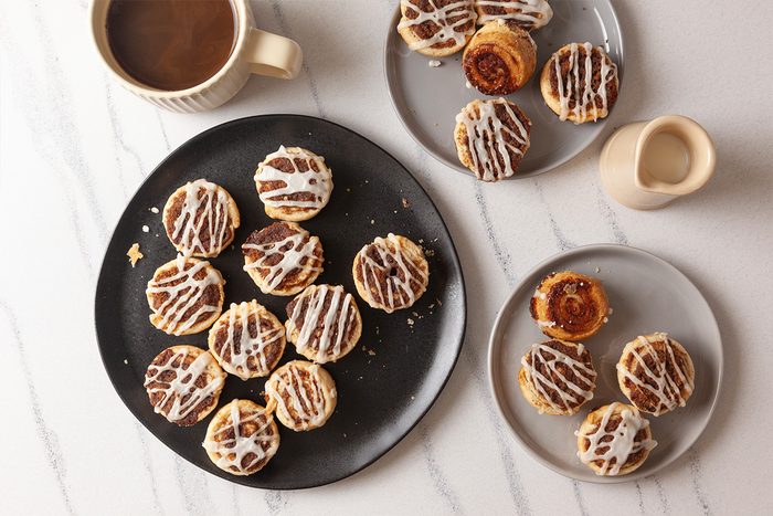 Overhead shot of Pie Crust Cinnamon Rolls drizzled with white icing on two plates, set beside a cup of coffee and a cream pitcher on a white marble surface