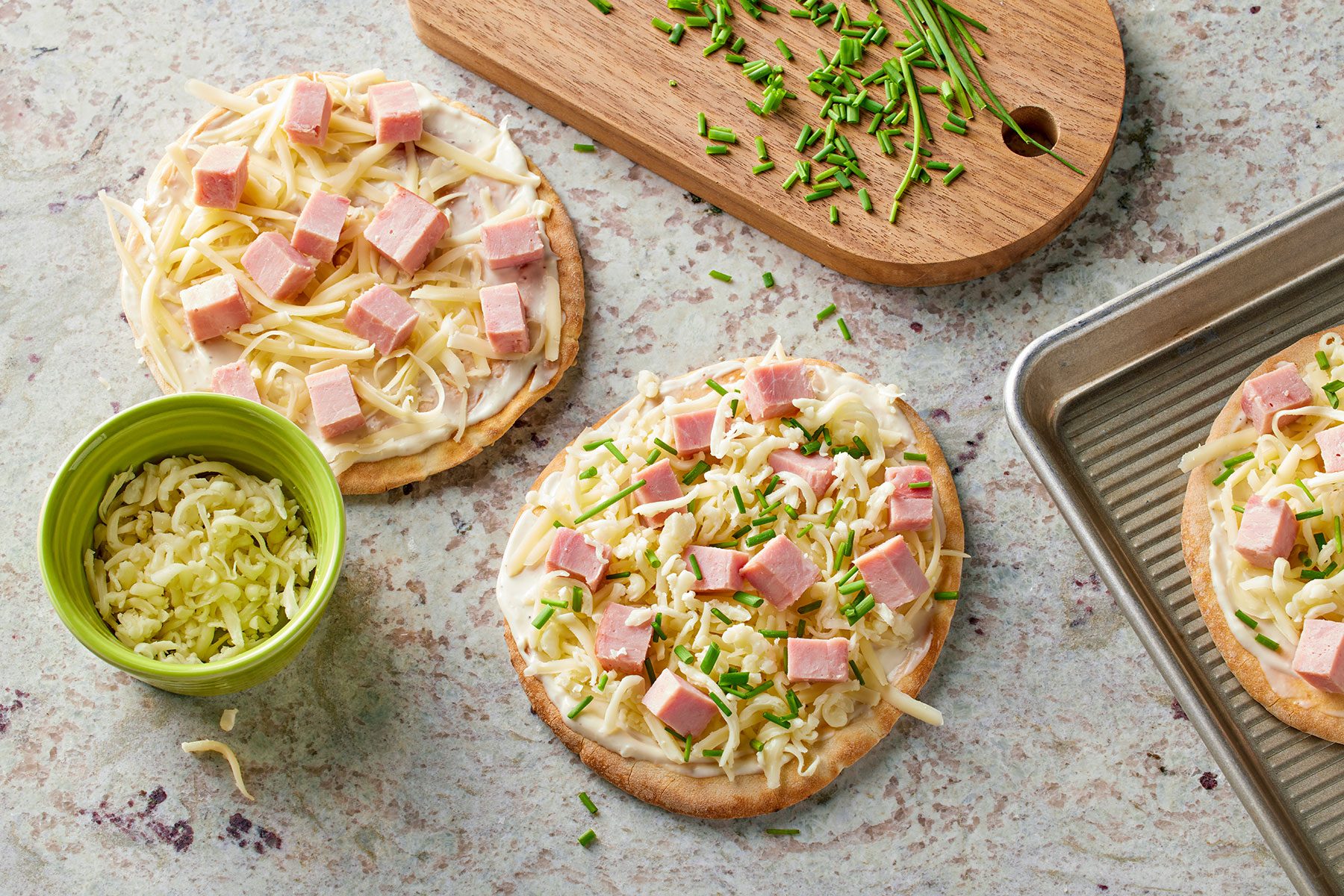 Topping pita bread with Swiss cheese, ham and mozzarella cheese, then sprinkling them with chives. cheese and chives on side.