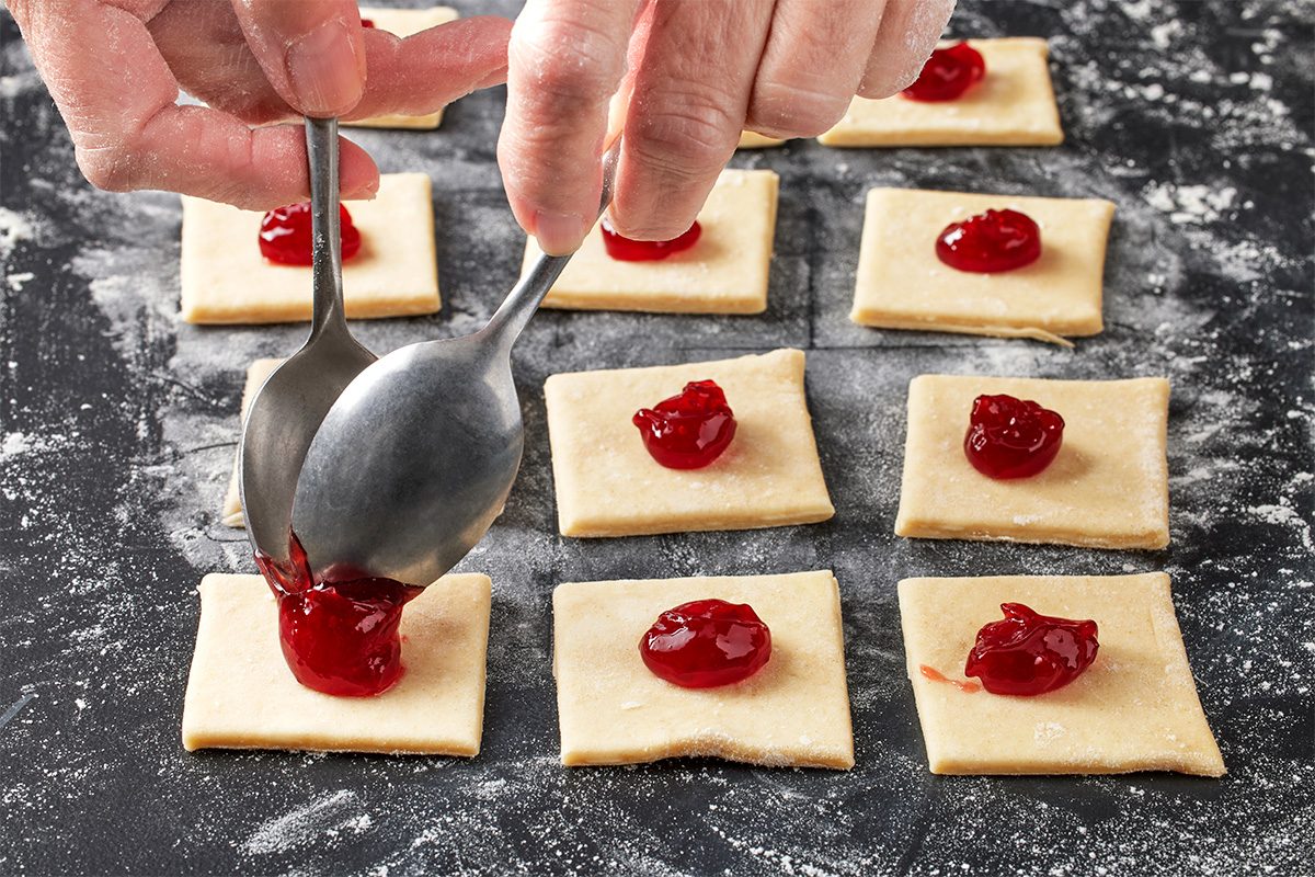 Hands using two spoons to place red cherry filling onto square pieces of raw dough arranged on a floured surface, preparing pastries or cookies.
