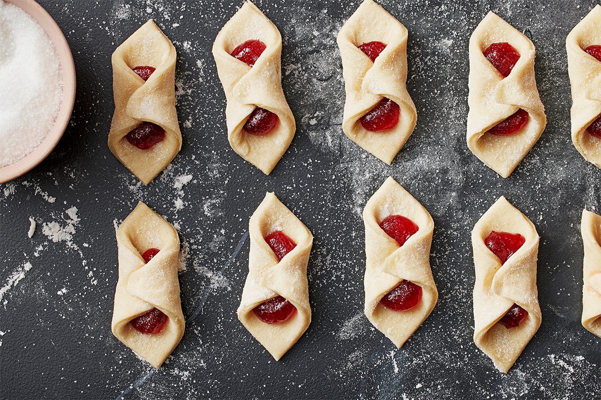 Unbaked kolaczki cookies filled with red fruit jam are arranged on a floured dark surface. A pink bowl of sugar sits to the side, and flour is scattered around the cookies.