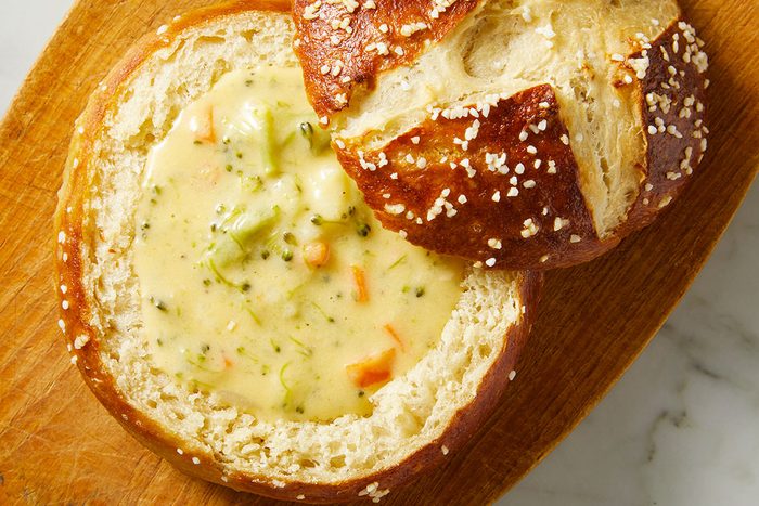 Overhead shot of a Pretzel Bread Bowl filled with creamy broccoli cheese soup, topped with carrots and broccoli, placed on a wooden board with the bread lid nearby.